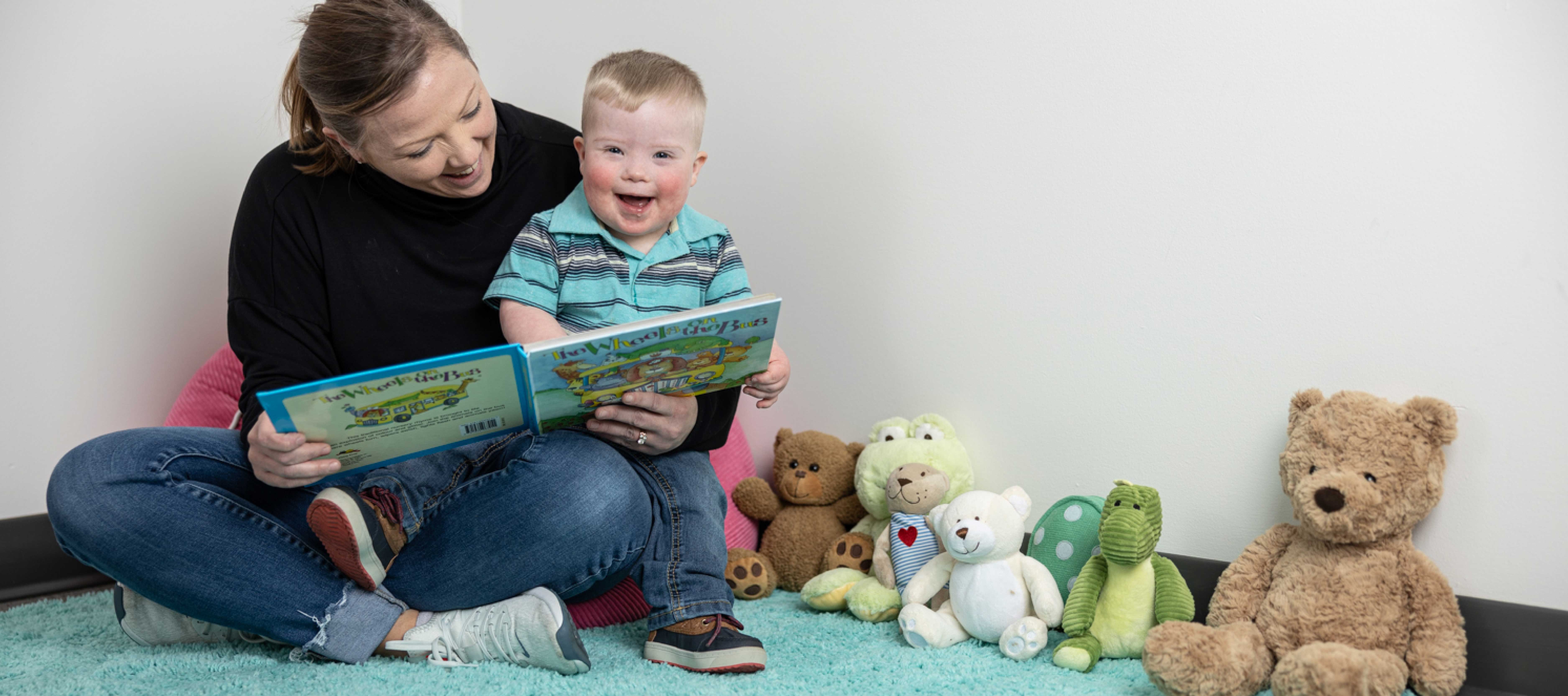 Toddler with Down syndrome on Mom’s lap, smiling while she reads