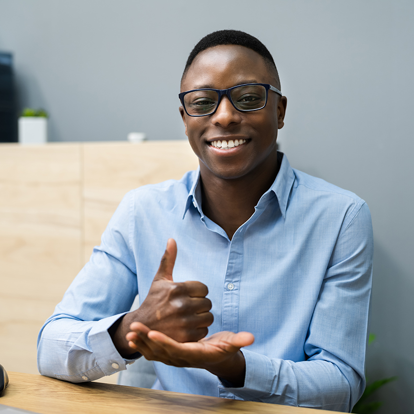 Young man seated at desk smiling at camera and using sign language